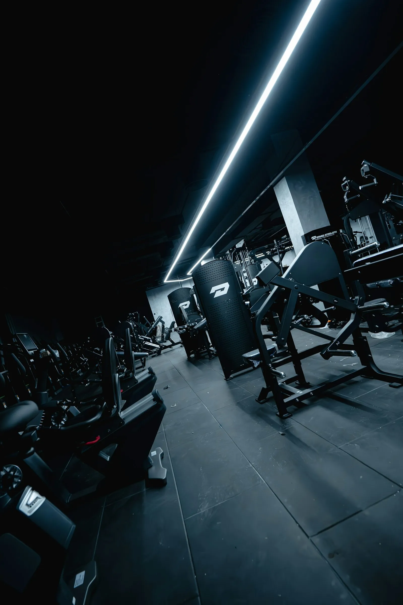 A wide view of an empty gym floor with machines and racks under strip lights